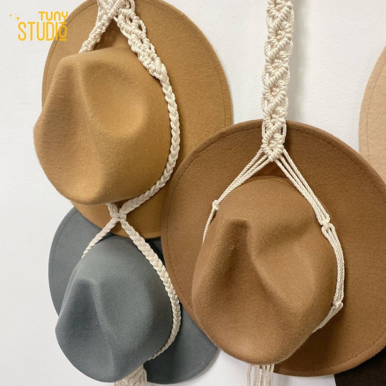 May include: Three brown felt hats with wide brims are hanging on a white wall using a white macrame rope hanger. The hats are arranged in a triangular shape.