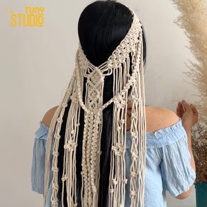 Peut inclure: Un accessoire pour cheveux en macramé de couleur crème, avec un bandeau et des mèches en cascade. Le design présente des nœuds complexes et des éléments décoratifs. L'image montre également le texte "TUNY STUDIO" en haut à gauche.