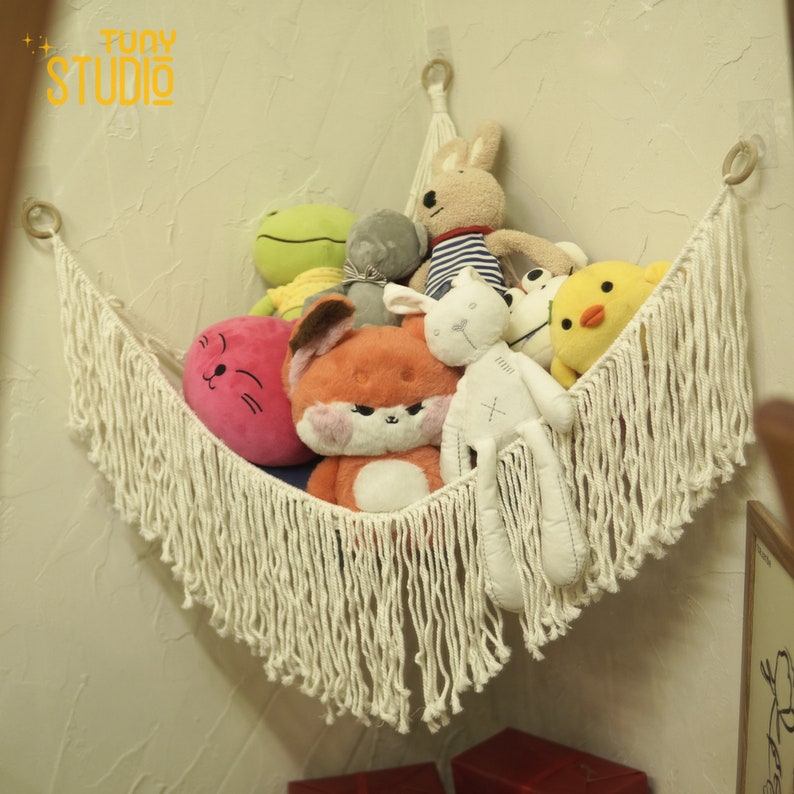May include: A white macrame wall hanging with a fringe border holds a collection of stuffed animals, including a pink cat, a red fox, a white bunny, a gray elephant, a green frog, a yellow chick, and a brown and white striped bunny.