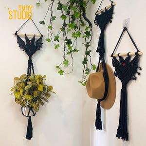 May include: Three black macrame wall hangings in the shape of bats with red eyes. The hangings are made with thick black cord and have wooden dowels at the top. The hangings are displayed on a white wall with a green plant hanging from one of them.