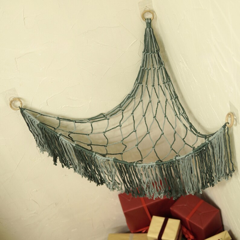 May include: A green and gray net hammock with fringe hanging from the bottom. The hammock is attached to the wall with two hooks. The hammock is empty.