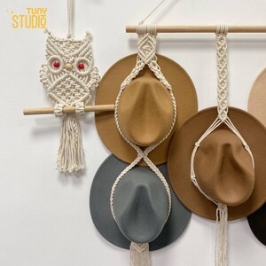 May include: A wall-mounted hat rack made of macrame with three hats hanging from it. The hats are brown, gray, and beige. The rack is made of white string and has a wooden dowel at the top. There is a white macrame owl hanging to the left of the rack.