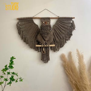 May include: A brown macrame owl wall hanging with wooden dowel and beads. The owl has large eyes and a detailed design.