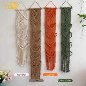 May include: Four macrame wall hangings in natural, latte, carrot, and olive green. Each has a wooden dowel at the top for hanging and multiple pockets. The natural and olive green hangings have a diamond pattern, while the latte and carrot have a chevron pattern.