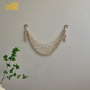 Op de afbeelding: Een witte macramé wandhanger met een netvormig ontwerp en kwastjes aan beide uiteinden. De hanger is bevestigd aan een witte muur.