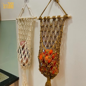 May include: Two macrame fruit holders hanging on a wall. One is off-white and filled with red apples, the other is brown and filled with red tomatoes. Both have wooden dowels and tassels.