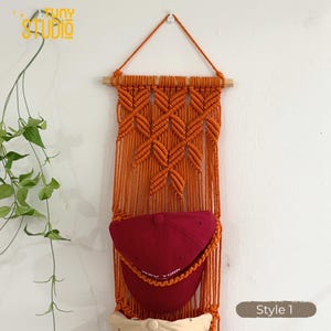 May include: An orange macrame wall hanging with a wooden dowel and a triangular hanger. The wall hanging has a diamond pattern and holds two baseball caps, one red and one cream-colored. The text "Style 1" is in the bottom right corner.
