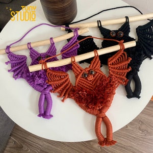 May include: Three macrame bat wall hangings with wooden dowels. The bats are in purple, orange, and black. Each bat has googly eyes.