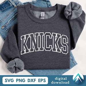 May include: A dark charcoal gray crewneck sweatshirt featuring the word KNICKS printed in a large white collegiate-style font across the chest. The garment has long sleeves with ribbed cuffs and a ribbed neckline with a visible brand label inside.