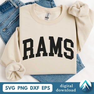 Peut inclure: Sweat-shirt crème avec le mot "RAMS" en lettres noires et grasses. Le sweat-shirt est plié sur un jean bleu. Le bas de l'image contient le texte "SVG PNG DXF EPS" et "téléchargement numérique".