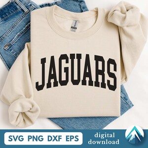Op de afbeelding: Crèmekleurige sweatshirt met het woord "JAGUARS" in vette, zwarte letters in collegestijl. De sweatshirt is opgevouwen op een blauwe spijkerbroek. De onderkant van de afbeelding bevat de tekst "SVG PNG DXF EPS" en "digitale download".