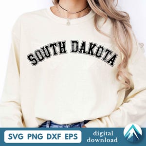 Puede incluir: Una sudadera de manga larga de color crema con el texto SOUTH DAKOTA impreso en una fuente negra, negrita y arqueada de estilo universitario en el pecho. La prenda tiene un cuello redondo y mangas largas, ofreciendo un aspecto sencillo y limpio.