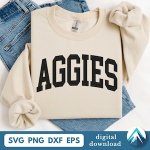 Op de afbeelding: Een crèmekleurige sweatshirt met het woord "AGGIES" in vette, zwarte blokletters. De sweatshirt is opgevouwen op een blauwe spijkerbroek. De onderkant van de afbeelding bevat de tekst "SVG PNG DXF EPS" en "digitale download".