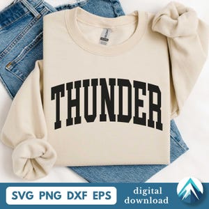 Puede incluir: Una sudadera de color crema con la palabra "THUNDER" en letras negras y en negrita, arqueadas. La sudadera está doblada sobre unos vaqueros azules. La parte inferior de la imagen incluye el texto "SVG PNG DXF EPS" y "digital download".