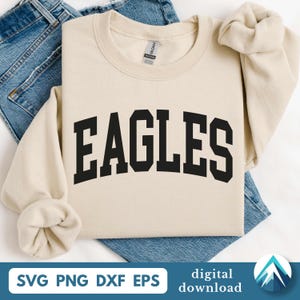 May include: A cream-colored sweatshirt with the word "EAGLES" in bold, black, block letters across the chest. The sweatshirt is folded on top of a pair of blue jeans. The bottom of the image has the text "SVG PNG DXF EPS" and "digital download".
