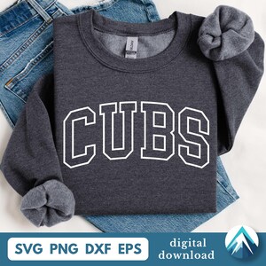 Puede incluir: Una sudadera gris oscuro con la palabra "CUBS" en letras blancas grandes. La sudadera está doblada sobre unos vaqueros azules. Las mangas están dobladas. La parte inferior de la imagen incluye el texto "SVG PNG DXF EPS digital download".