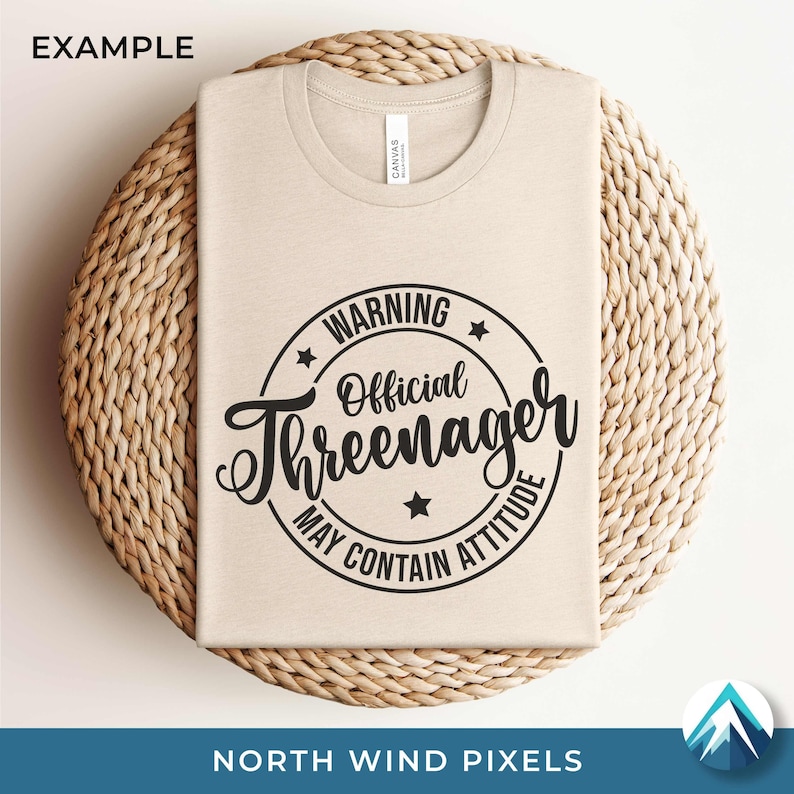 K&ouml;nnte beinhalten: Beiges T-Shirt mit Rundhalsausschnitt und schwarzem, kreisf&ouml;rmigem Aufdruck mit der Aufschrift "WARNING Official Threenager MAY CONTAIN ATTITUDE". Das Shirt liegt auf einem runden, gewebten Platzdeckchen.