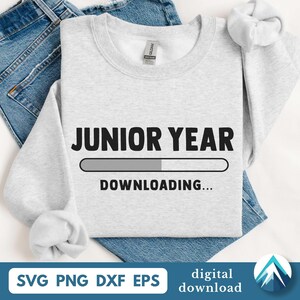 以下が含まれることがあります： 「JUNIOR YEAR」の文字とローディングバーのグラフィック、そして「DOWNLOADING...」の文字がプリントされたライトグレーのスウェットシャツ。スウェットシャツは青いデニムジーンズの上に折りたたまれています。