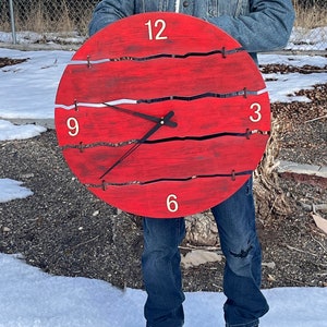 May include: A red wooden clock with black hands and white numbers. The clock face is cracked and has a distressed look. The clock is being held by a person wearing a denim jacket.