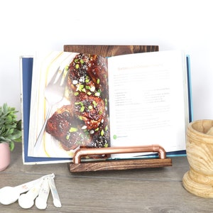May include: A wooden cookbook stand with a copper pipe book holder. The stand is holding a cookbook open to a recipe for Barbecued Chicken Thighs. The stand is made of dark stained wood and is on a wooden surface.