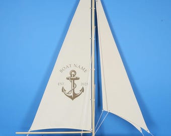 May include: A model sailboat with a dark wood hull and a cream-colored sail. The sail features a brown graphic of an anchor with the text BOAT NAME and EST. 2023. The boat is mounted on a rectangular wooden base with a vertical support pole.