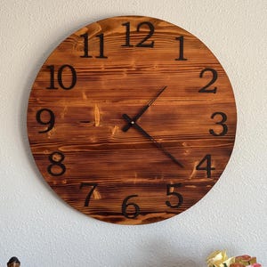 May include: A round wooden wall clock with a dark brown finish. The clock face features black numerals and hands. The clock is mounted on a white wall. The clock's diameter is approximately 12 inches.