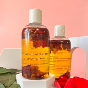 May include: Two bottles of Vanilla Rose Body Oil with white lids, containing a golden liquid and rose petals. The label says "Kandaka Collections" and "Helps moisturize and nourish skin". The bottles are on a white platform.