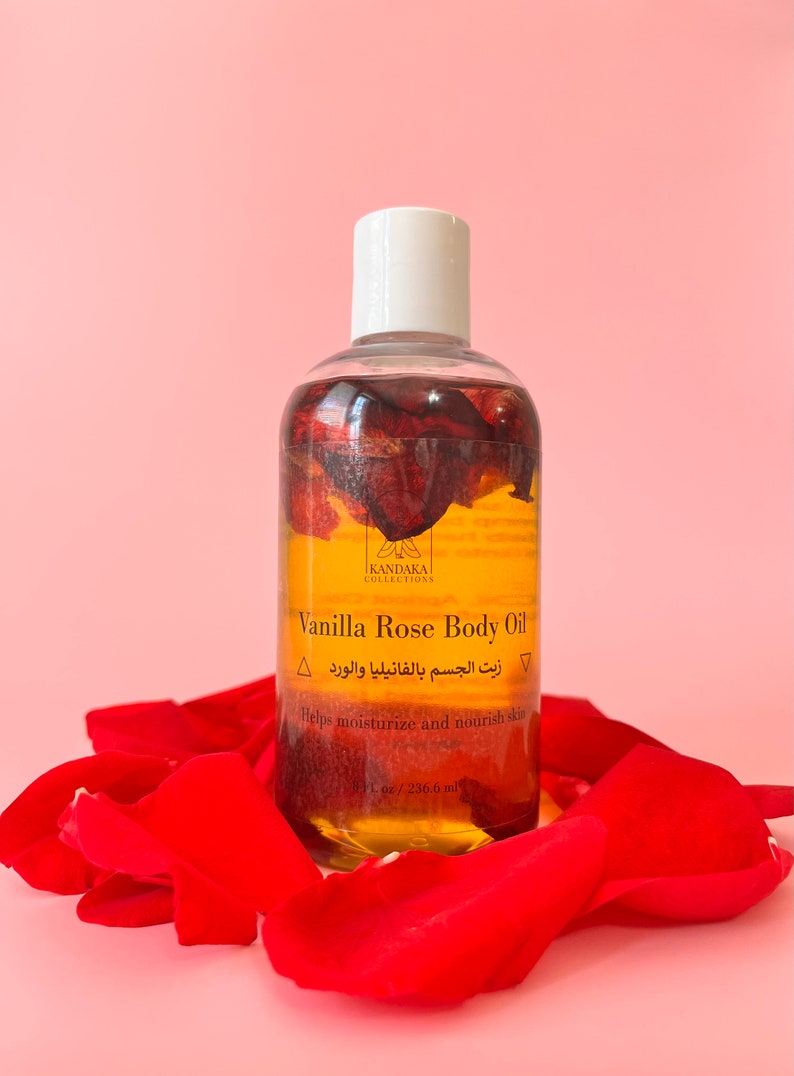 Puede incluir: Una botella de vidrio transparente de aceite corporal de vainilla y rosa con una tapa blanca, rodeada de p&eacute;talos de rosa rojos. La botella contiene un l&iacute;quido dorado con p&eacute;talos de rosa en su interior. El texto en la botella dice "Vanilla Rose Body Oil" y "236,6 ml".