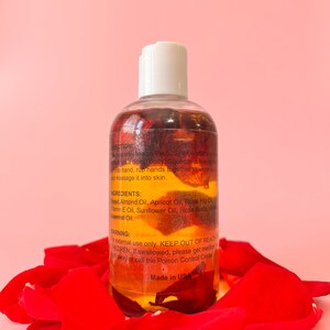 Puede incluir: Una botella transparente de aceite corporal con tapa blanca, llena de p&eacute;talos de rosas rojas y l&iacute;quido amarillo. La botella est&aacute; rodeada de p&eacute;talos de rosas rojas. Texto en la botella incluye ingredientes e instrucciones de uso. Hecho en EE. UU.