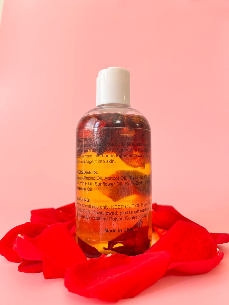 Puede incluir: Una botella transparente de aceite corporal con tapa blanca, llena de p&eacute;talos de rosas rojas y l&iacute;quido amarillo. La botella est&aacute; rodeada de p&eacute;talos de rosas rojas. Texto en la botella incluye ingredientes e instrucciones de uso. Hecho en EE. UU.