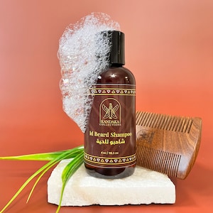 May include: A brown bottle of beard shampoo with a white foamy lather on top. The bottle is labeled "Id Beard Shampoo" and "شامبو اللحية" in Arabic. The bottle is sitting on a white stone surface with a wooden comb next to it.