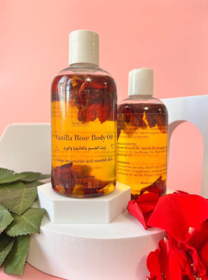 Puede incluir: Dos botellas transparentes de aceite corporal de vainilla y rosa con tapas blancas, llenas de l&iacute;quido dorado y p&eacute;talos de rosa. La etiqueta dice "Kandaka Collections" y "Helps moisturize and nourish skin". Las botellas est&aacute;n sobre una plataforma blanca.