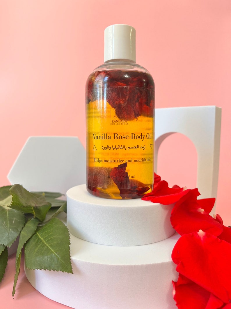 Puede incluir: Una botella transparente de aceite corporal de vainilla y rosa con tapa blanca, llena de p&eacute;talos de rosa secos. La etiqueta dice "Vanilla Rose Body Oil" y "Helps moisturize and nourish skin." La botella es de 236 ml. El producto se exhibe sobre un pedestal blanco con p&eacute;talos de rosa rojos.