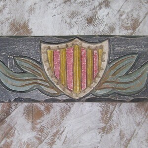 May include: A wooden wall plaque featuring a carved shield design. The shield is painted in red, yellow and white stripes. The shield is flanked by two carved leaves.