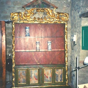 May include: An antique wooden cabinet with a red interior and gold trim. The cabinet has three shelves holding decorative ceramic mugs. The bottom section features painted panels. The cabinet is in a room with stone walls.