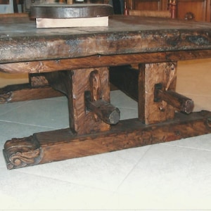 Può includere: Un grande tavolo in legno rustico con un piano spesso di colore marrone scuro e gambe intagliate intricate. Il tavolo è realizzato in legno riciclato e ha un aspetto unico e artigianale.