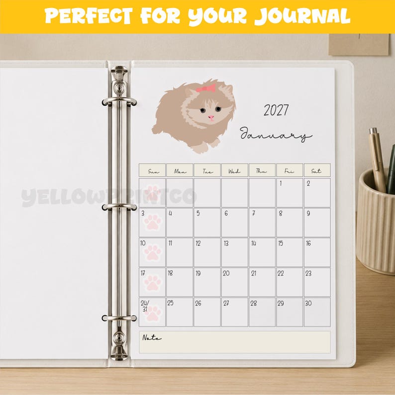 May include: A white binder containing a January 2027 calendar page. The page features a light brown cat illustration with a pink bow, the text January 2027, and a grid with light pink paw prints marking the first column of dates for the month.