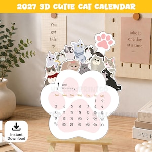 May include: A 2027 3D cutie cat calendar shaped like a large white paw print with pink pads. The calendar features various illustrated cats at the top and displays the month of January 2027 with black text on a white background, including a pink paw icon.