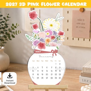 May include: A 2027 desk calendar shaped like a vase filled with colorful watercolor flowers. The calendar displays the month of January with a grid of dates. A pink ribbon graphic is printed across the neck of the vase. The design is white with pink, yellow, and purple floral illustrations.