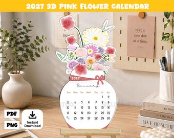 May include: A 2027 desk calendar shaped like a vase filled with colorful watercolor flowers. The calendar displays the month of January with a grid of dates. A pink ribbon graphic is printed across the neck of the vase. The design is white with pink, yellow, and purple floral illustrations.