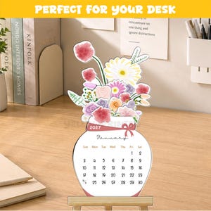 May include: A 2027 desk calendar shaped like a vase filled with colorful flowers. The calendar displays the month of January with dates from 1 to 30. It features a pink ribbon graphic around the neck of the vase and sits on a small wooden stand.