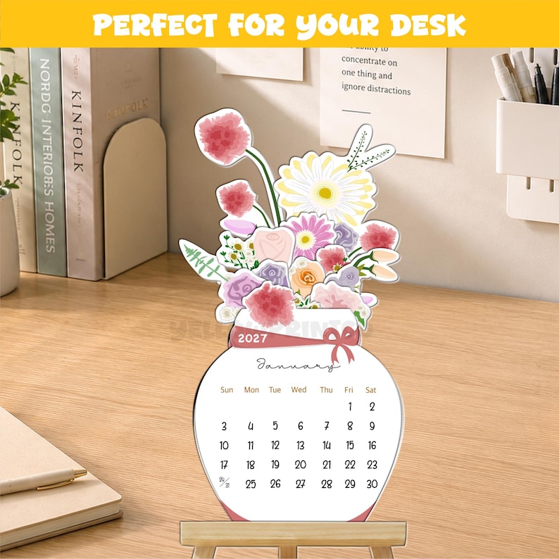 May include: A 2027 desk calendar shaped like a vase filled with colorful flowers. The calendar displays the month of January with dates from 1 to 30. It features a pink ribbon graphic around the neck of the vase and sits on a small wooden stand.