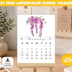 May include: A 2027 mini watercolor floral calendar featuring a white background with a purple wisteria illustration at the top. The calendar displays the month of January with black text and numbers, presented on a light wooden easel stand.