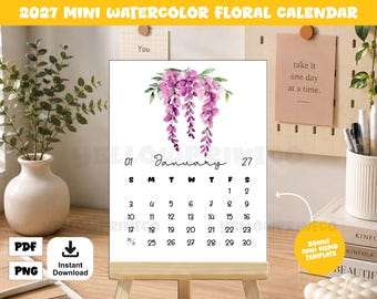 May include: A 2027 mini watercolor floral calendar featuring a white background with a purple wisteria illustration at the top. The calendar displays the month of January with black text and numbers, presented on a light wooden easel stand.