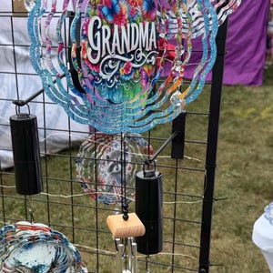 May include: A circular wind spinner featuring a blue glittery scalloped edge and a central design of colorful flowers, butterflies, and the word GRANDMA in white script. Below it hangs a wind chime with a wooden block and silver metal tubes.