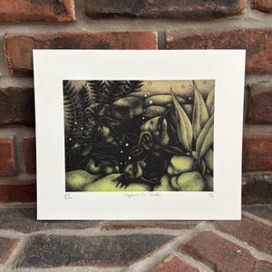 May include: Black and yellow print titled "Caymans On Biale" featuring a detailed illustration of several caymans in a swampy environment. The artwork is set against a white background and framed, with the artist's initials and date visible.