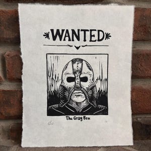 May include: Black and white linocut print on textured paper. The print features a stylized image of a masked figure with the word "WANTED" at the top. Below the image is the text "The Gray Fox".