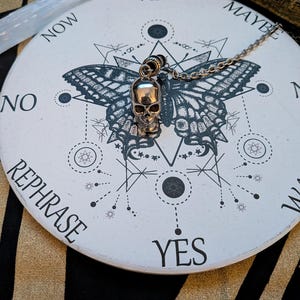 May include: A silver-toned skull pendant on a metal chain resting on a circular white board. The board features a black butterfly graphic, geometric lines, and the words YES, MAYBE, NO, and REPHRASE printed in a black sans-serif font.
