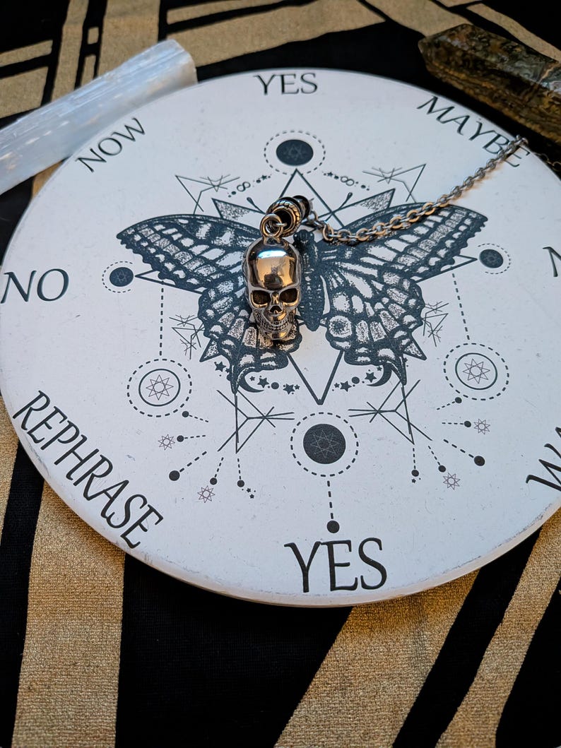 May include: A silver-toned skull pendant on a metal chain resting on a circular white board. The board features a black butterfly graphic, geometric lines, and the words YES, MAYBE, NO, and REPHRASE printed in a black sans-serif font.
