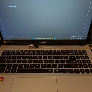 May include: A silver-toned skull pendant attached to a delicate metal chain, resting on the keyboard of a silver Acer laptop. The laptop screen displays a document with the text Unlock Your Spiritual Gifts and Thank you.